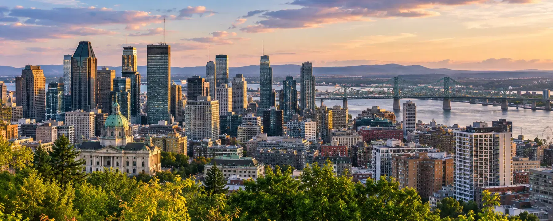 A city skyline at sunset seen from a wooded hillside, with a green-domed cathedral below.